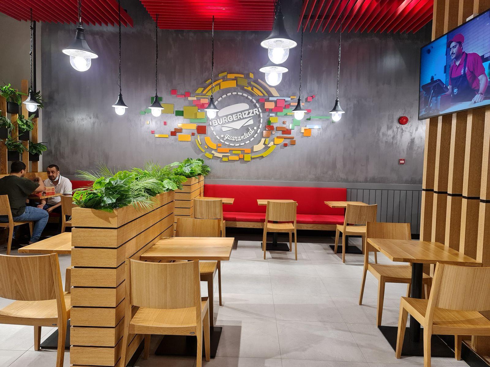 Burgerizzr, Makkah: timber tables beside a planter, facing the red banquette and logo wall